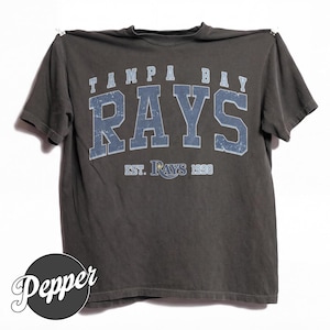 May include: A charcoal gray short-sleeve t-shirt featuring the text TAMPA BAY RAYS in a distressed blue font across the chest. Below the main text, the shirt displays EST. RAYS 1998 in a smaller blue font. A circular black logo with the word Pepper is visible.
