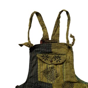 May include: Close-up of a patchwork overall with adjustable straps. The overall features a pocket with a button closure and floral and geometric patterns in shades of green, black, and yellow. The straps are striped.