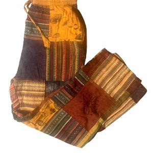 May include: Patchwork pants in shades of brown, yellow, and blue. The pants feature a variety of patterns, including stripes, floral designs, and mushroom graphics. The waist has an elastic band and drawstring.