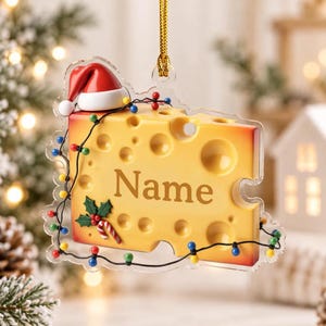 May include: A yellow Swiss cheese-shaped Christmas ornament featuring a red Santa hat, a string of colorful holiday lights, and a holly sprig. The word Name is printed in brown letters across the center of the cheese block.