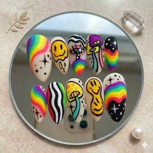 May include: A set of ten long, almond-shaped artificial nails with colorful, psychedelic designs. The designs include rainbows, smiley faces, mushrooms, and abstract patterns. The nails are arranged on a round mirror.