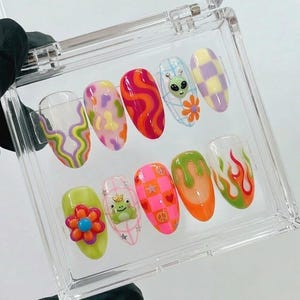 May include: A set of ten artificial nails in a clear plastic case. The nails feature various designs, including swirls, flowers, an alien, a frog, and flames. Colors include orange, pink, green, yellow, and purple.