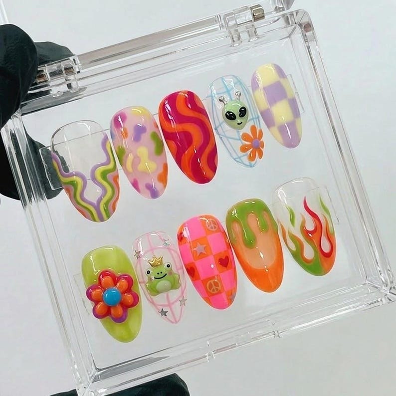 May include: A set of ten artificial nails in a clear plastic case. The nails feature various designs, including swirls, flowers, an alien, a frog, and flames. Colors include orange, pink, green, yellow, and purple.