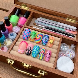 May include: A wooden box filled with nail art supplies. The box contains colorful nail polish bottles, glitter pots, nail files, brushes, and a variety of decorated false nails. The nails feature designs like checkered patterns, aliens, and smiley faces.