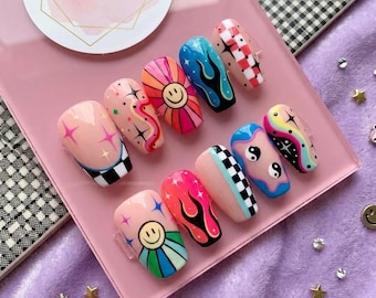 May include: A collection of colorful, press-on nails with various designs. The nails feature smiley faces, flames, stars, and checkered patterns in shades of pink, blue, orange, and red. The nails are arranged on a pink surface.