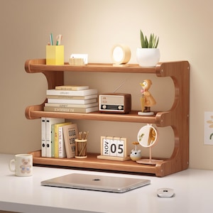 May include: A three-tiered wooden desktop organizer shelf featuring a curved side design. The shelves hold books, a vintage-style radio, a duck figurine, a calendar showing 05, a small mirror, a pencil holder, and a potted plant on the top shelf.