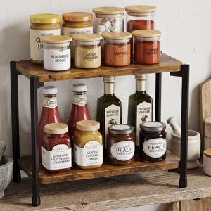 May include: A two-tier countertop spice rack with a dark wood-grain finish and black metal frame. The shelves hold various glass jars with bamboo lids, including labels for powdered sugar, garlic powder, paprika, cayenne pepper, and fruit spreads.