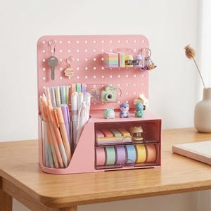 May include: A pink desk organizer featuring a pegboard back panel with hanging hooks, a clear pen holder filled with pastel pens, and two transparent drawers containing colorful sticky notes, washi tape rolls, and small animal-shaped figurines.