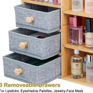 May include: A light wood cosmetic organizer featuring three removable grey fabric drawers with round wooden knobs, positioned next to open shelves holding various skincare and makeup bottles. The text 3 Removable Drawers For Lipsticks, Eyeshadow Palettes, Jewelry, Face Mask is visible at the bottom.