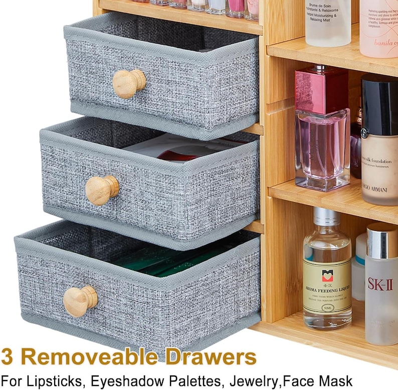 May include: A light wood cosmetic organizer featuring three removable grey fabric drawers with round wooden knobs, positioned next to open shelves holding various skincare and makeup bottles. The text 3 Removable Drawers For Lipsticks, Eyeshadow Palettes, Jewelry, Face Mask is visible at the bottom.