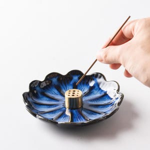Puede incluir: Un porta incienso de cerámica azul y blanca con forma de flor. Tiene un cilindro de latón con agujeros en el centro. Un palito de incienso se sostiene sobre el soporte. El soporte mide aproximadamente 15 cm de diámetro.