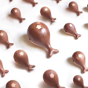 May include: Wooden whale-shaped decorative objects in various sizes. Each whale is a rich brown color with a lighter-colored flower inlay on top. The whales have a smooth finish and are arranged on a white background.