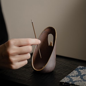 Pode incluir: Um porta-incenso de cerâmica marrom com um design curvo único. O suporte tem uma pequena abertura e um detalhe dourado. Um incenso é colocado no suporte. O suporte está sobre uma superfície escura.