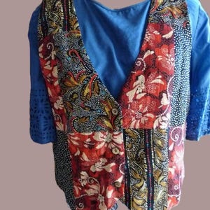 Op de afbeelding: Een blauw shirt onder een mouwloos vest met een patchworkontwerp van rode, zwarte en witte bloemen- en abstracte patronen. Het vest heeft drie knopen aan de voorzijde en het blauwe shirt heeft korte mouwen met uitgesneden details.