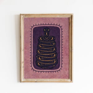 May include: A framed wall art print featuring a dark purple central panel with a coiled snake illustration. The snake is decorated with gold crescent moons and dots. The background is a textured dusty rose colour with a dark purple border frame.