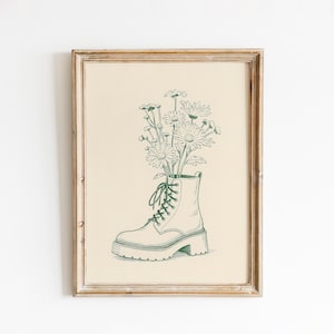 May include: A framed art print showing a green line drawing of a combat boot filled with daisies. The boot has a chunky sole and is laced up. The daisies have multiple petals and are in full bloom. The background is a light beige colour.