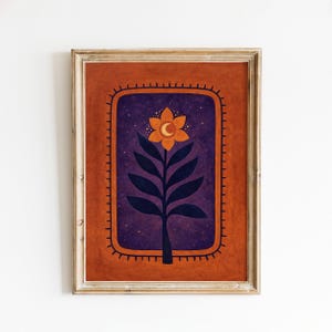 May include: A framed wall art print featuring a central orange flower with a crescent moon center, set against a dark purple starry background. The design is enclosed in a rectangular border with small radiating lines, all set on a terracotta background.