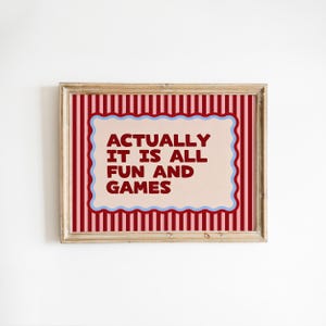 May include: A rectangular framed print featuring a light pink background with a dark red and light pink vertical striped border. In the center, a scalloped light blue frame surrounds the dark red text: ACTUALLY IT IS ALL FUN AND GAMES.