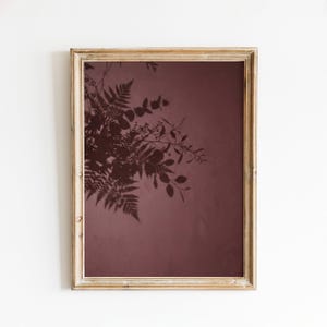 May include: A rectangular wooden frame containing a print with a dark maroon background. The print features a dark silhouette of fern leaves and eucalyptus branches extending from the upper left corner across the solid colored surface.