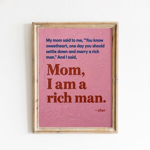 Może przedstawiać: Oprawiony wydruk artystyczny z teksturowanym różowym tłem i złotą ramą. Wydruk zawiera cytat "Mom, I am a rich man" w ciemnoczerwonym tekście, przypisany Cher. Górny tekst jest niebieski.