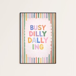 May include: A framed wall art print featuring a background of vertical multicolored stripes in shades of blue, green, yellow, orange, and pink. Centered is a scalloped-edge white rectangle containing the text BUSY DILLY DALLY ING in colorful letters.