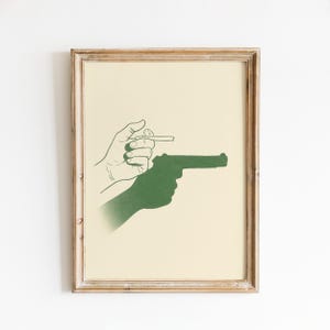 Peut inclure: Impression encadrée représentant une silhouette verte d'un pistolet et d'une main tenant une cigarette. L'œuvre est posée sur un fond crème et encadrée dans un cadre en bois brun clair. L'œuvre d'art a un style graphique minimaliste.