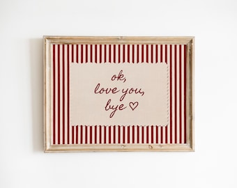 May include: A framed artwork with a red and white striped background. The artwork features a rectangular beige fabric with the words "ok, love you, bye" in burgundy script, and a heart symbol. The frame is made of light-coloured wood.