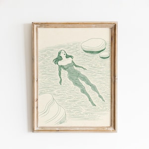 Op de afbeelding: Een ingelijste kunstdruk met een groene illustratie van een persoon die in het water zwemt. De persoon wordt getoond met lang haar en een badpak. Twee groene stenen drijven in het water. Het kunstwerk is ingelijst in een lichtbruine houten lijst.