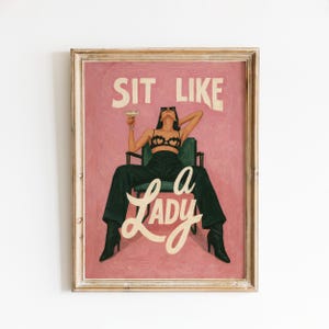 Può includere: Stampa artistica incorniciata con una donna con occhiali da sole, seduta su una sedia verde, che tiene in mano un cocktail. Lo sfondo rosa ha le parole "SIT LIKE" sopra e "A LADY" sotto. L'opera d'arte è in una cornice di legno.