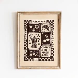 Könnte beinhalten: Ein gerahmter Druck mit einem dunkelbraunen Grafikdesign auf hellbeigem Hintergrund. Das Kunstwerk zeigt eine Mokkakanne, Kaffeetassen, ein Croissant, Kirschen und den Text "COFFEE FIRST, MAGIC LATER", umgeben von einem karierten Rand.