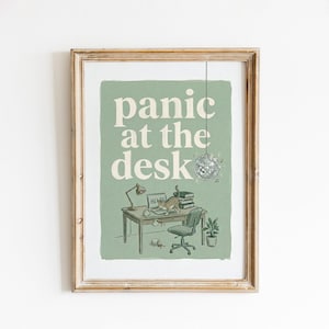 May include: A framed wall art print featuring a sage green background with the text "panic at the desk" in white. The illustration shows a desk with a lamp, a laptop, a stack of books, a potted plant, a disco ball, and a cat standing on the desk.