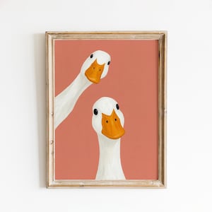May include: A framed art print showing two white geese with orange beaks and black eyes against a coral background. The geese are arranged in a quirky, off-centre composition, creating a playful and engaging visual.