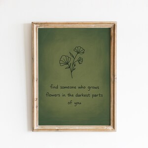Könnte beinhalten: Gerahmter Kunstdruck mit grünem Hintergrund und einfacher Blumenillustration. Der Text lautet: "Finde jemanden, der Blumen in den dunkelsten Teilen von dir wachsen lässt." Der Rahmen ist hellbraun.