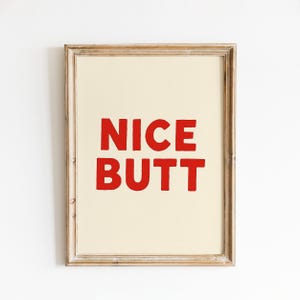 May include: A framed print with the words "NICE BUTT" in bold red letters. The text is centered on a cream-colored background, set within a light brown wooden frame. The artwork is a humorous piece.