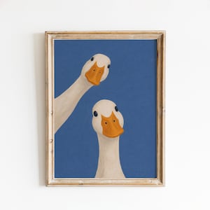 May include: Framed art print featuring two white geese with orange beaks and black eyes against a blue background. The geese are positioned in a whimsical, slightly askew manner, creating a playful and engaging visual.