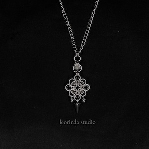 Peut inclure: Un collier à chaîne argentée doté d'un pendentif en métal complexe et tissé avec un motif central carré. Le pendentif est orné de petits anneaux circulaires et d'une pointe conique unique dirigée vers le bas. Le texte leorinda studio est visible.