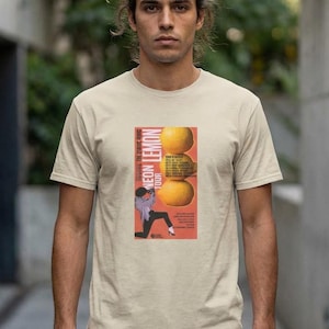 Könnte beinhalten: Beiges T-Shirt mit einem Aufdruck "Neon Lemon Tour". Die Grafik zeigt drei Zitronen und eine Person im schwarzen Anzug. Das T-Shirt ist kurzärmlig und hat einen Rundhalsausschnitt.