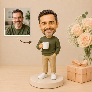 May include: A figurine of a man holding a white mug, wearing a green sweater, khaki pants, and white sneakers. The figurine is on a round, beige base. A small gift box with a bow and a bouquet of roses are in the background.