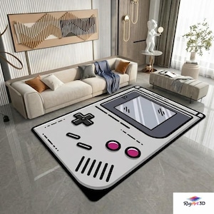 Puede incluir: Una alfombra rectangular grande con un diseño de mando de videojuego retro. La alfombra presenta un fondo gris con contornos y detalles negros, incluyendo una cruceta, botones y una pantalla. Dos círculos rosas están en la parte inferior derecha.