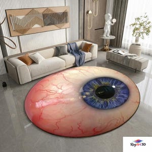Puede incluir: Una alfombra redonda con un diseño de ojo realista. La alfombra presenta un iris azul, una pupila negra y una esclerótica roja y rosa con venas visibles. La alfombra está colocada sobre un suelo de baldosas grises en un salón.