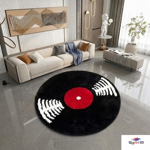 Puede incluir: Una alfombra redonda negra con un centro rojo y un diseño gráfico blanco, que se asemeja a un disco de vinilo. La alfombra está colocada sobre un suelo de baldosas grises en un salón, con un sofá beige y arte mural.