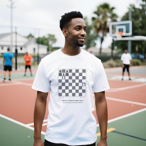 Puede incluir: Camiseta blanca con un diseño gráfico de tablero de ajedrez y el texto "WHITE TO MOVE! MATE IN 2!". El diseño del tablero de ajedrez es en blanco y negro. La camiseta se lleva al aire libre.
