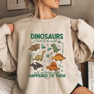 May include: A beige crewneck sweatshirt featuring a graphic print with the text "DINOSAURS didn't do math and we know what HAPPENED TO THEM". The design includes illustrations of five different dinosaurs, mathematical symbols, and geometric shapes.
