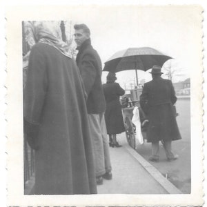 May include: A black and white photo of four people standing in a line. The person in the front is wearing a long coat and a headscarf. The person behind them is wearing a dark coat and pants. The person behind them is wearing a dress and the person in the back is wearing a coat and pants. There is a cart with a white cloth and a black umbrella in the background.