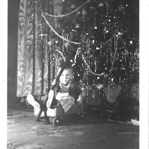 May include: A black and white photo of a child sitting in a wagon in front of a Christmas tree decorated with white tinsel.