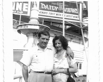 May include: A black and white photo of a couple standing in front of a fountain. The man is wearing a white shirt and the woman is wearing a floral dress. The sign behind them reads "DAILY NEWS" and "ALWAYS FIRST WITH THE LATEST NEWS".