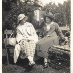 May include: Three people are sitting on a bench outdoors. The person in the middle is wearing a white hat and a plaid skirt. The person on the right is wearing a dark hat and a long coat. The person on the left is wearing a dark hat and a suit.