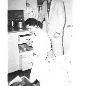 May include: A black and white photo of a woman in a white blouse looking into a cabinet drawer. A man in a suit jacket stands behind her.
