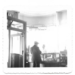 May include: A black and white photo of a man wearing a hat standing in front of a counter with the word "EXIT" above the door.