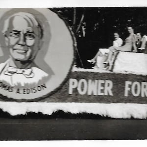 May include: A black and white photo of a parade float with a large portrait of Thomas A. Edison. The float is decorated with flowers and the text "Power for Defense".